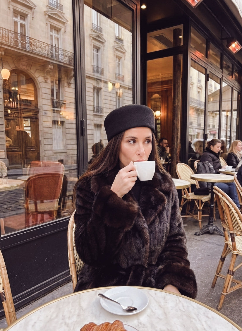 Woman in a fur coat and black hat drinking coffee in a Parisian cafe.