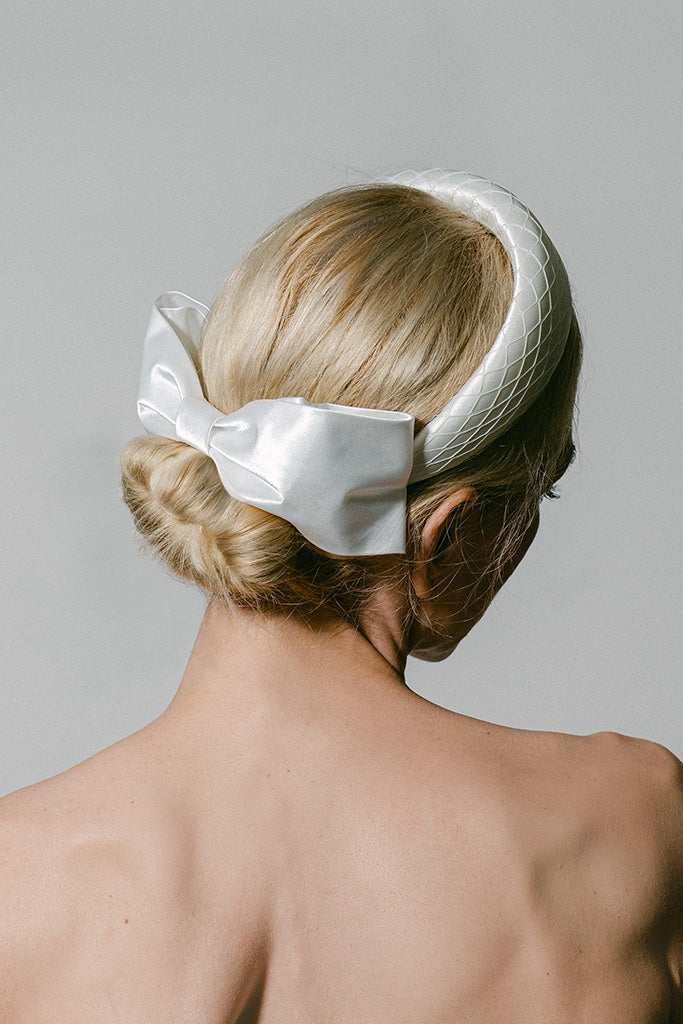 A blonde woman shown from the back wearing a white structured headband with a large satin bow, against a plain light gray studio background.