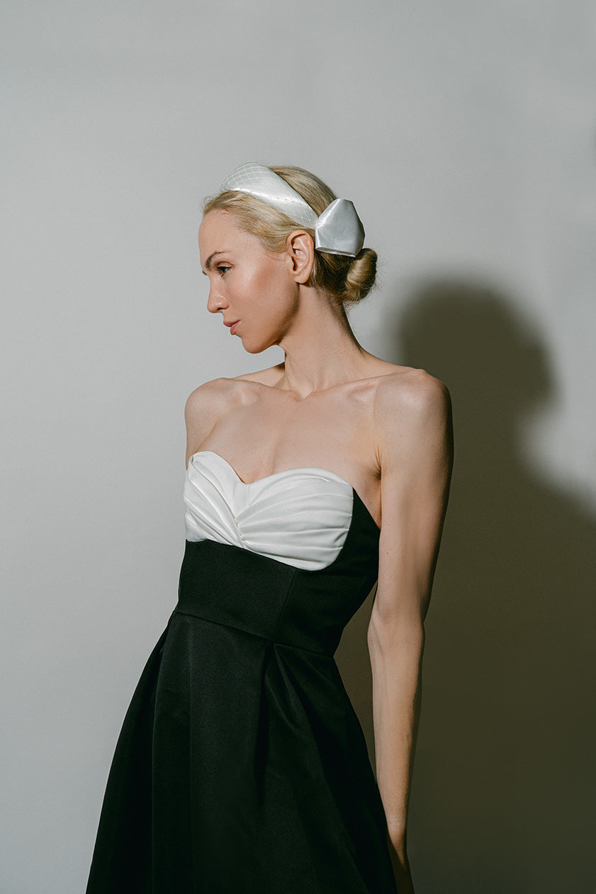 A woman in profile wearing a strapless black dress with a white sweetheart bodice and a white headband, photographed against a plain light gray studio background.