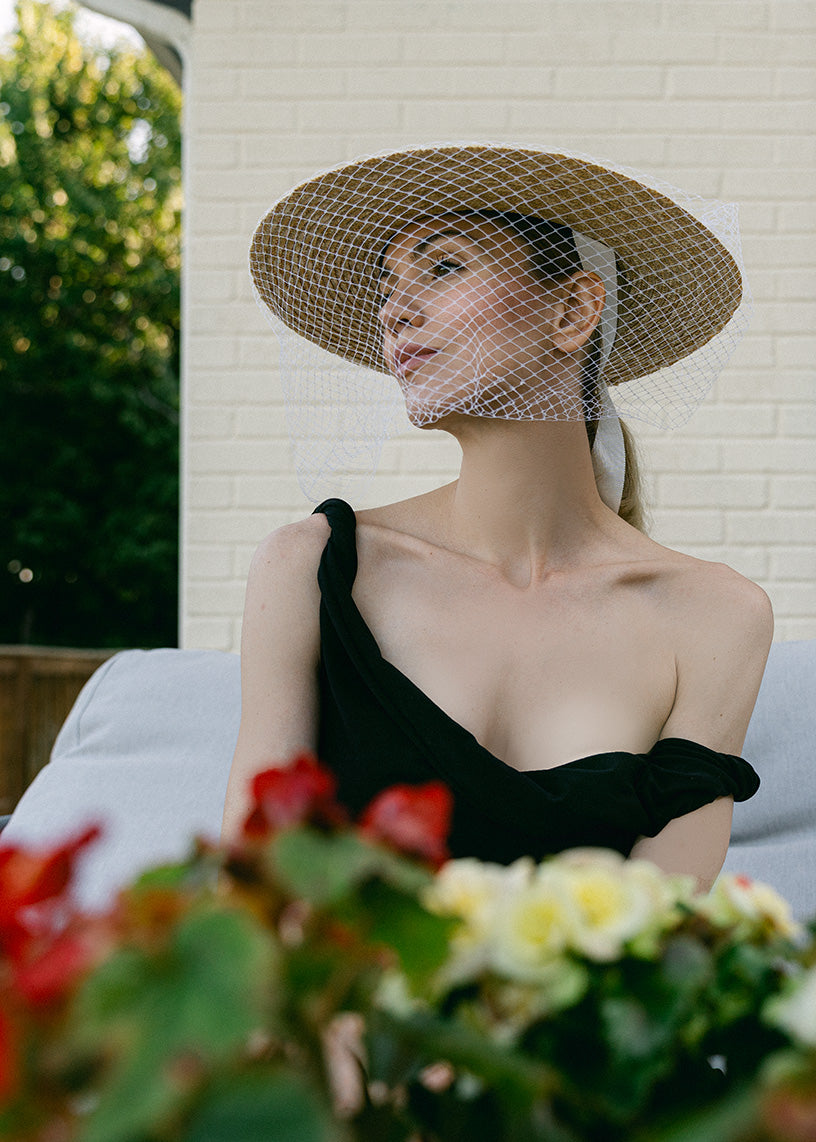 A woman wearing a wide-brimmed straw hat with a white mesh veil and a black one-shoulder dress, seated outdoors against a white brick wall with blurred flowers in the foreground.